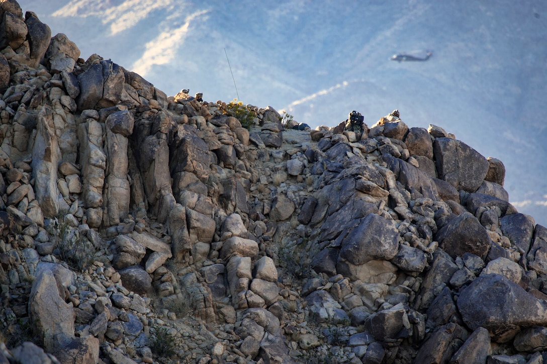 An Army AH-64 Apache Guard flies past U.S. Army soldiers in the Mojave Desert mountains while staking out a simulated battlefield during a rotation at Ft. Irwin, California’s National Training Center, Feb. 23, 2018. During the month-long rotation, 93d Air Ground Operations Wing units embedded with approximately 4,000 soldiers in the largest force-on-force live-fire exercise in the world. The 93d AGOW provided tactical air control party support to enhance interoperability for major combat operations downrange. (U.S. Air Force photo by Senior Airman Greg Nash)