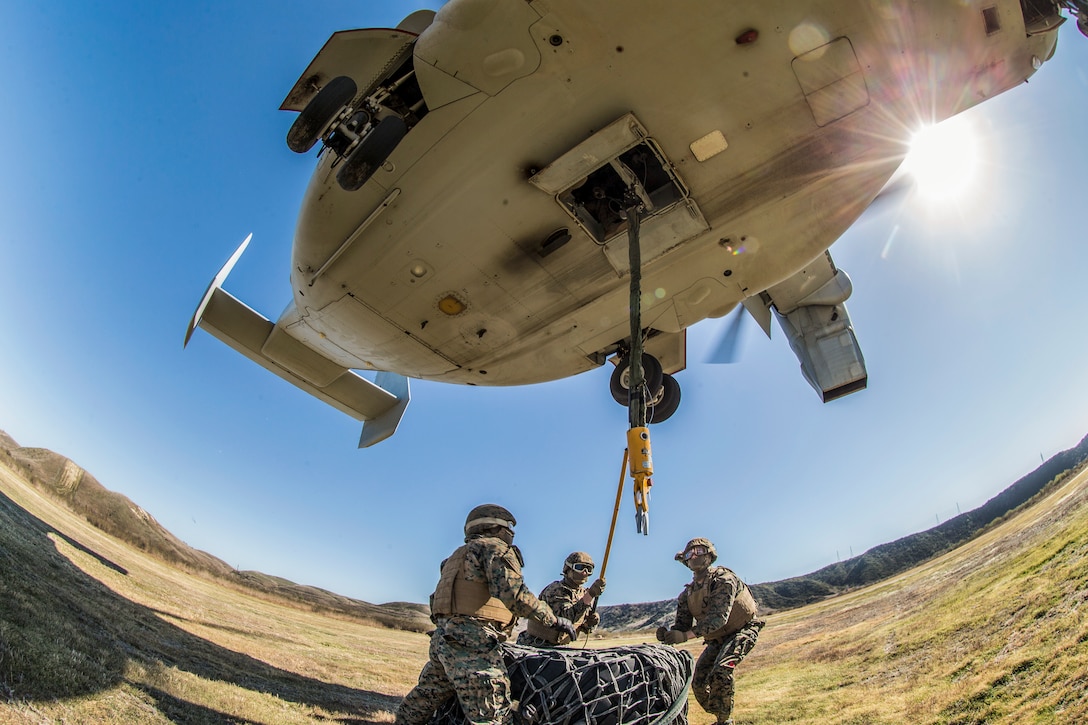 Marines attach a simulated load onto a helicopter hovering over them.