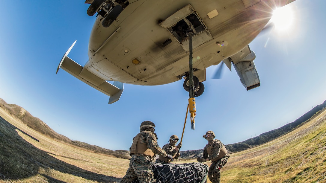 Marines attached a simulated load onto a helicopter hovering over them.