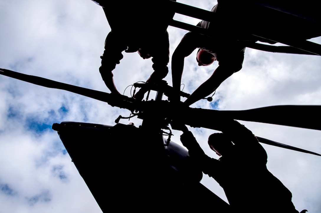 Airmen, shown in silhouette, perform maintenance on a helicopter's rotor against a cloudy sky.