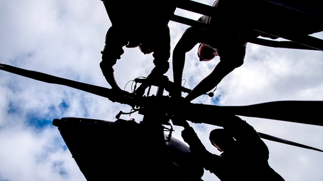 Airmen, shown in silhouette, perform maintenance on a helicopter's rotor against a cloudy sky.