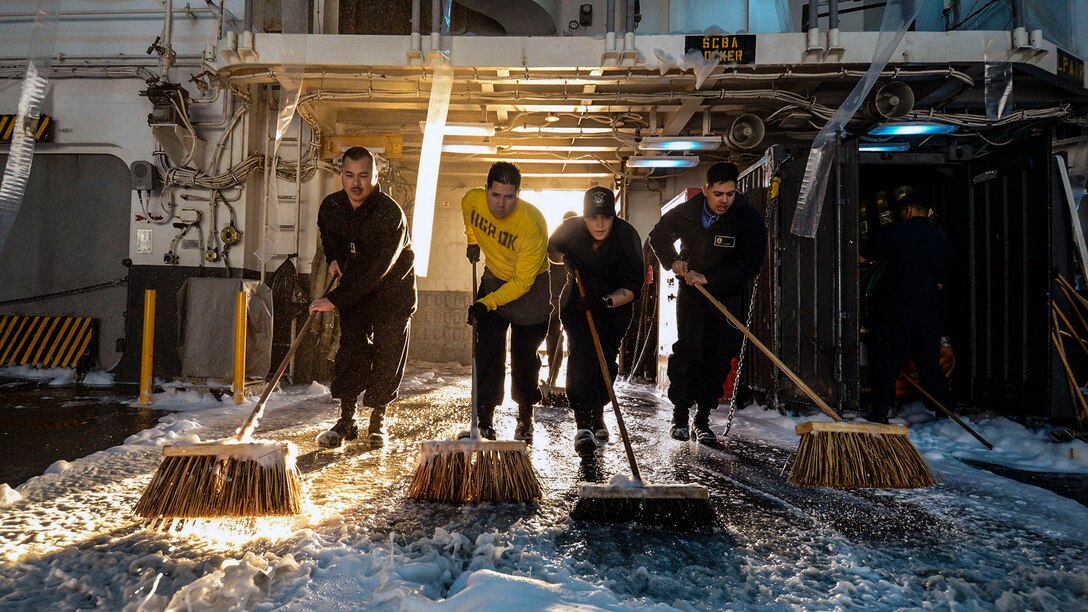 A row of sailors use large brooms to scrub the deck.