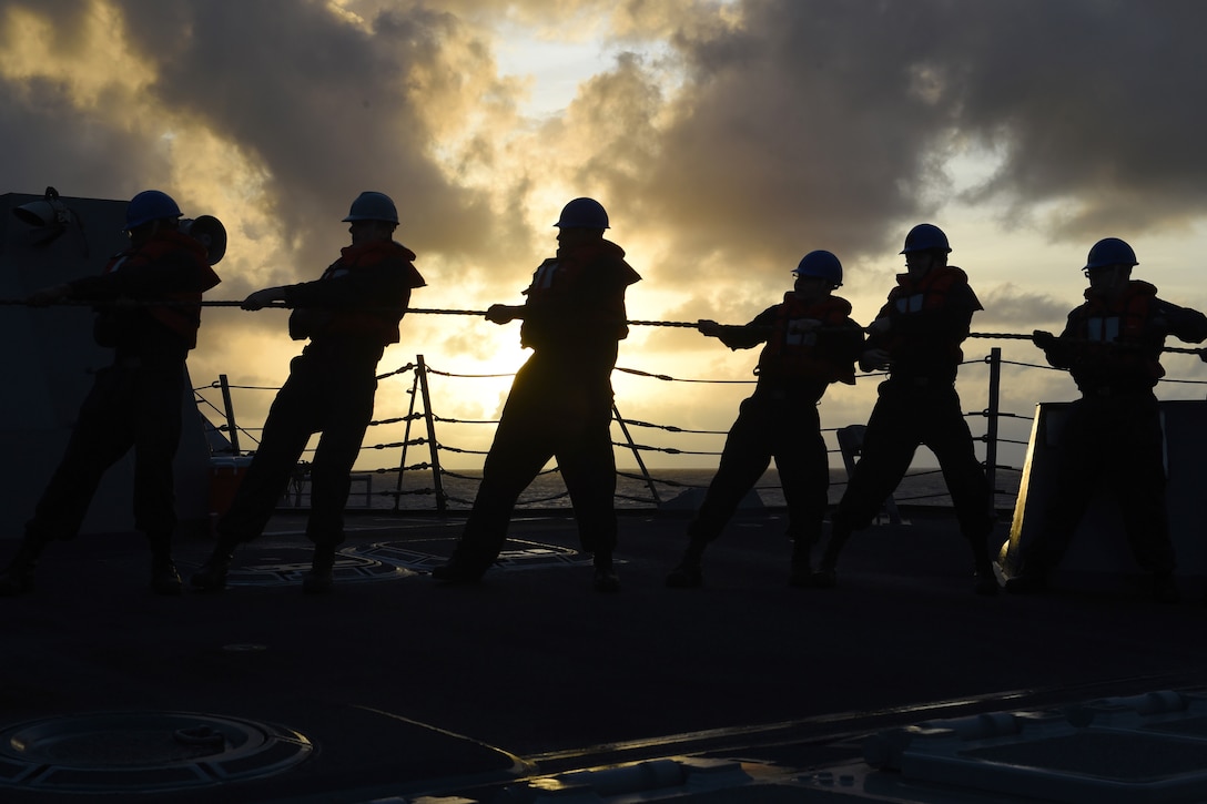 A line of sailors pull a line with colorful clouds in the background.