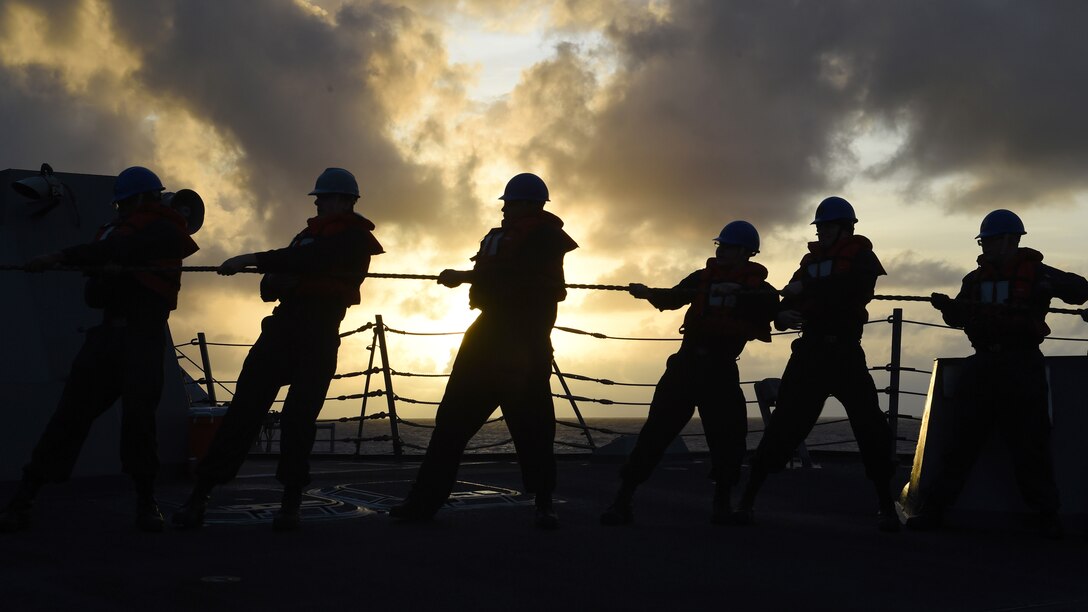 A line of sailors pull a line with colorful clouds in the background.