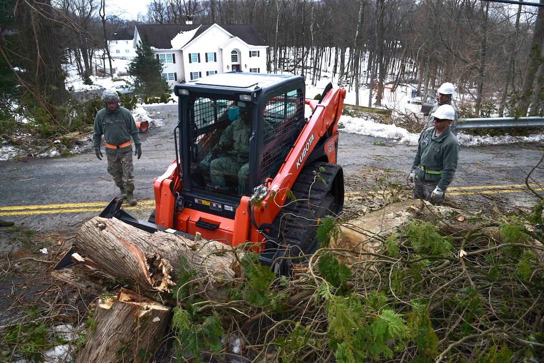 An airman uses skid-steer loader to remove branches and debris from a road.