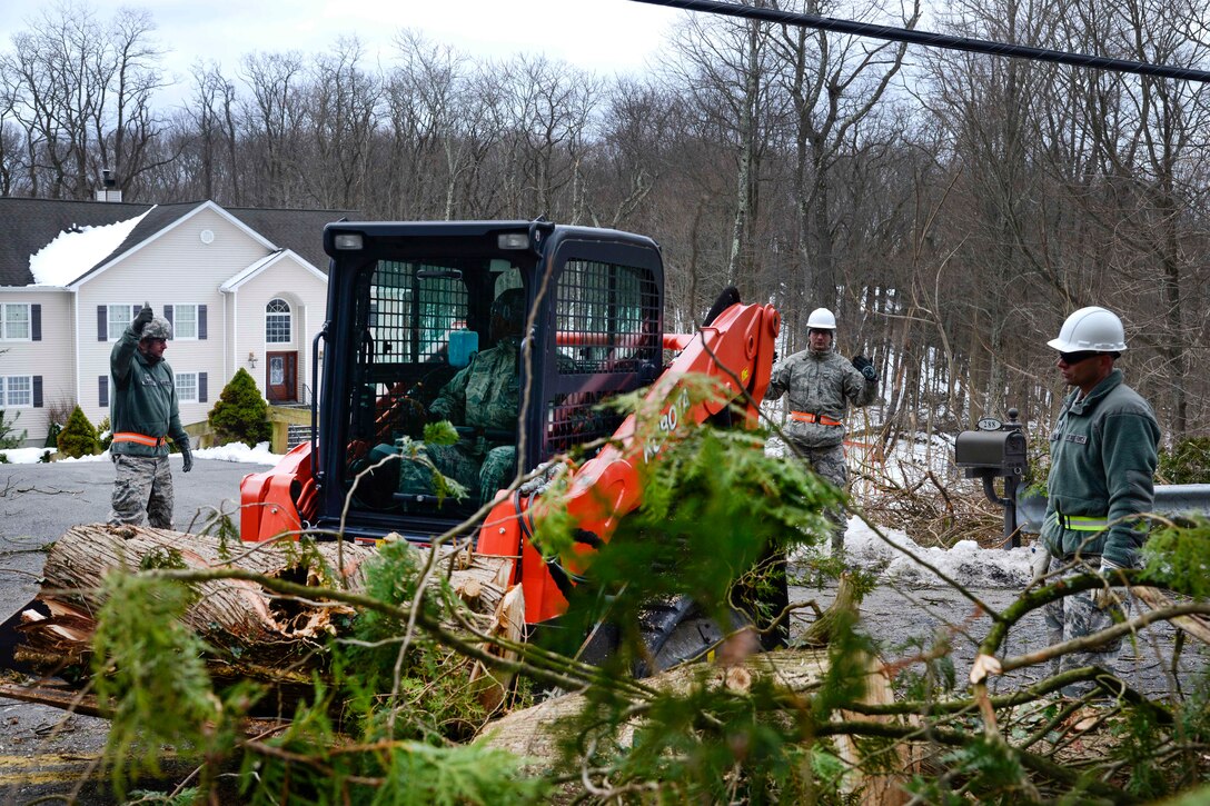 An airman operates a skid-steer loader to remove branches and debris from a road.
