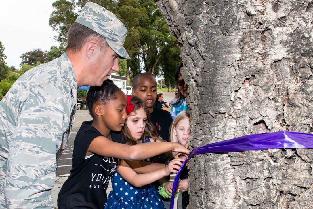 An airman helps children tie a ribbon around a tree.