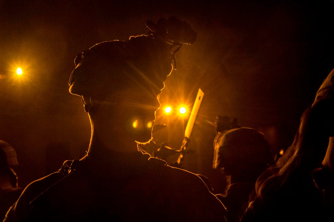 A Marine, shown in silhouette, listens to a night briefing as yellow lights shine.