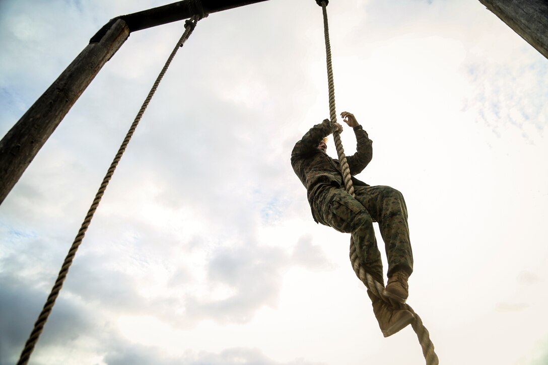 A Marine climbs a rope against a partly cloudy sky during an obstacle course.