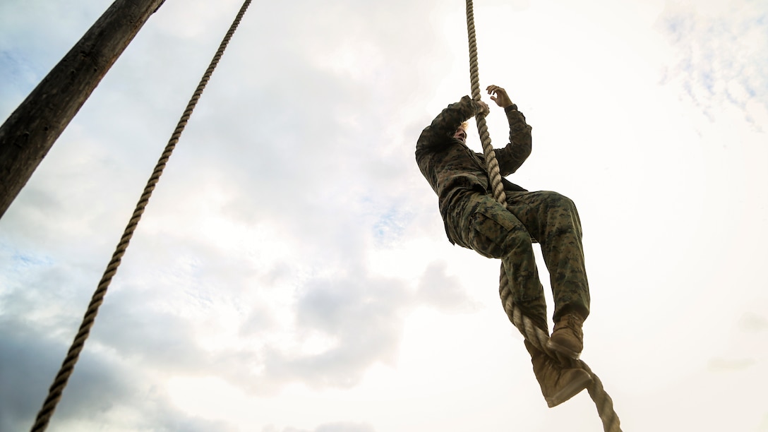 A Marine climbs a rope against a partly cloudy sky during an obstacle course.