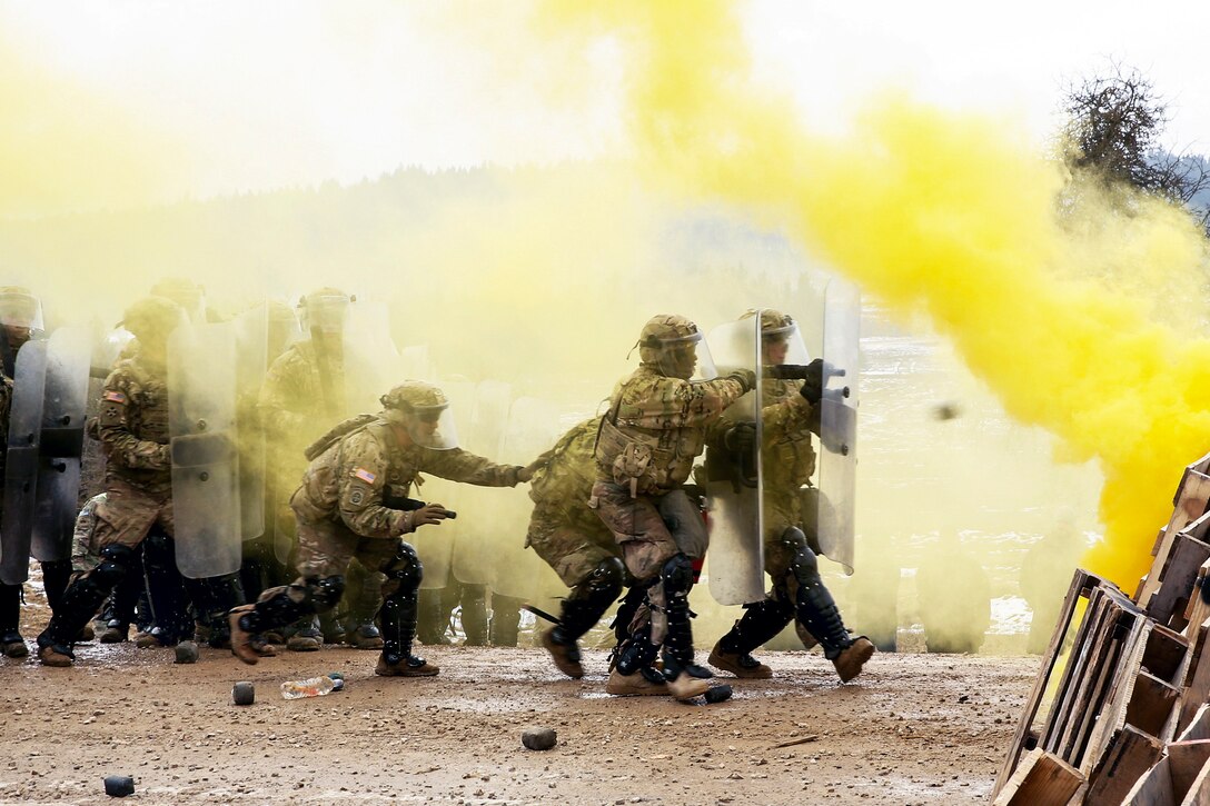 Soldiers conduct riot control training with shields as yellow smoke rises.