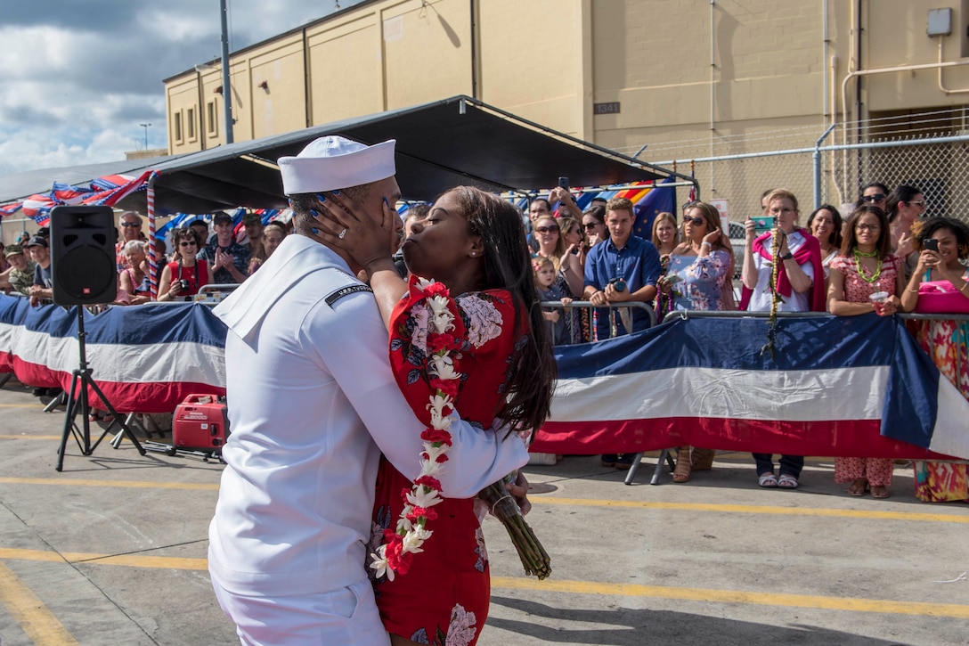 A sailor returning from a deployment embraces a loved one holding a lei as a crowd watches.