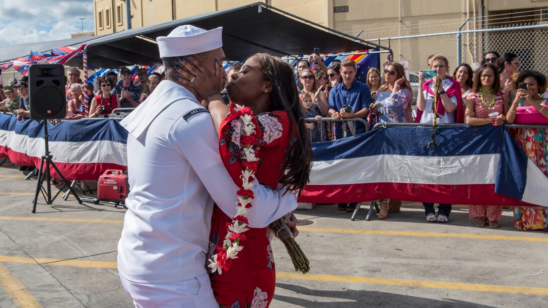 A sailor embraces a loved one holding a lei on a pier as a crowd watches.