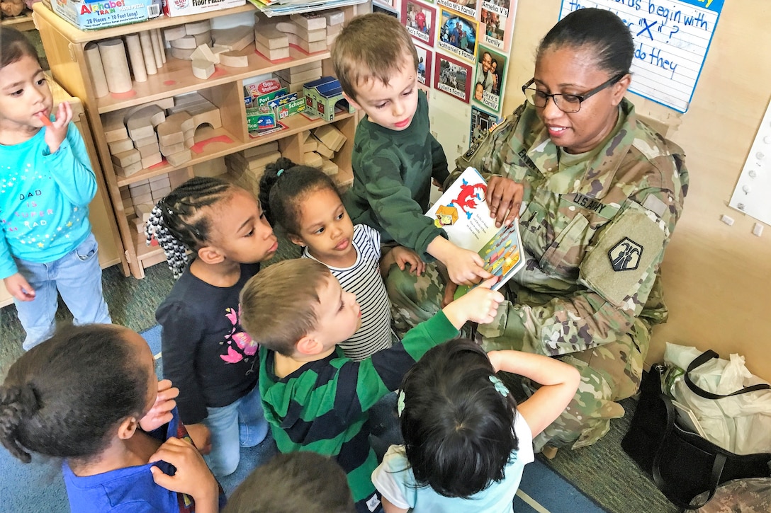 Children gather around a soldier reading to them, and study and touch the book she's holding.