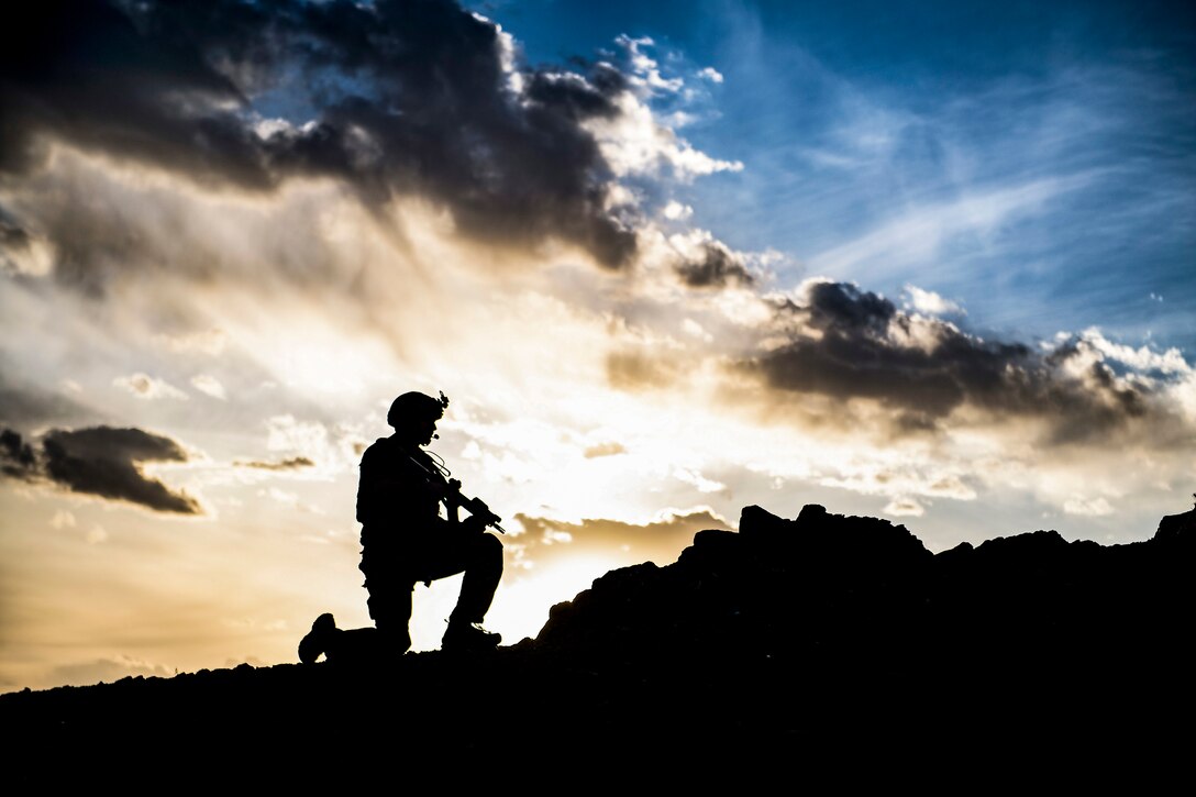 An airman, shown in silhouette, kneels with a weapon on rocky terrain, against a cloud- and light-streaked blue sky.