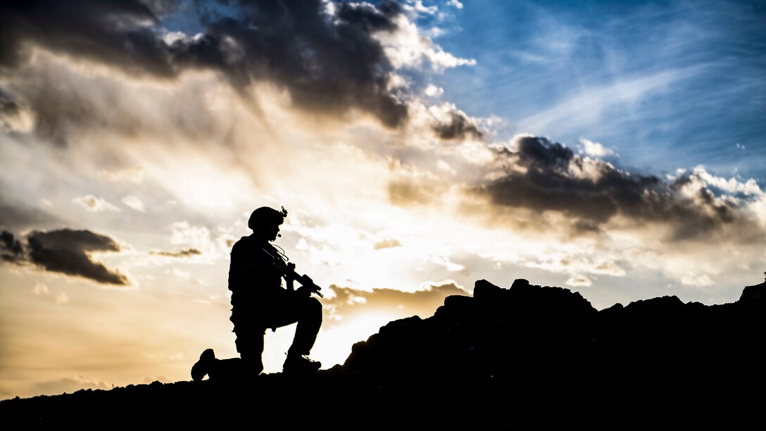 An airman, shown in silhouette, kneels with a weapon on rocky terrain, against a cloud- and light-streaked blue sky.