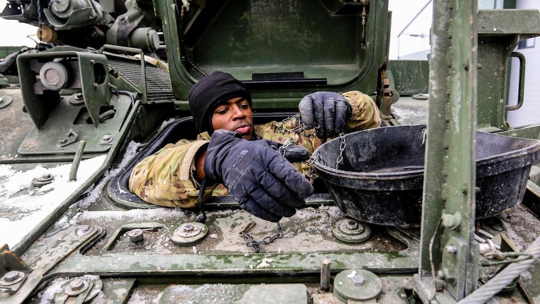 A soldier works on parts on top of a vehicle's exterior while perched partway outside the top opening of the vehicle.