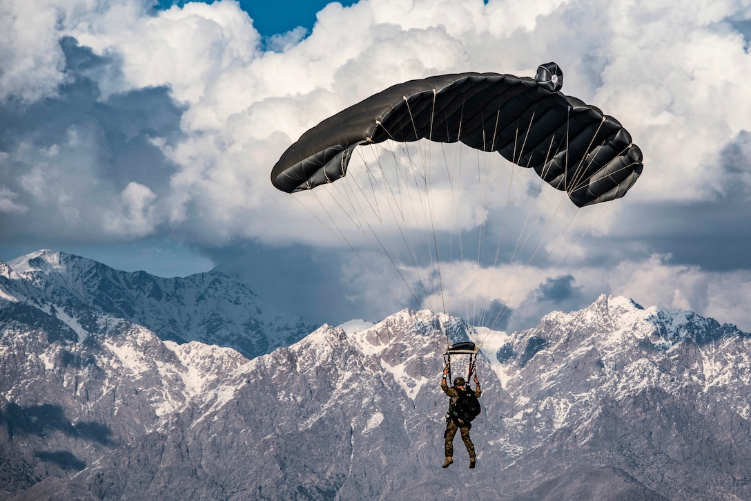 An airman parachutes against a backdrop of snow-capped mountain peaks and cloud-filled blue sky.