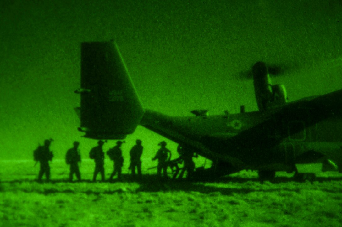 Soldiers, seen in silhouette in green light from a night vision device, board the back of an aircraft parked on flat terrain.
