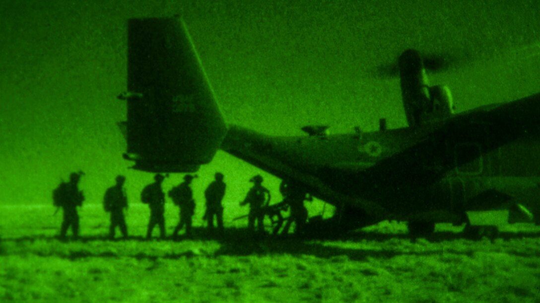 Soldiers, seen in silhouette in green light from a night vision device, board the back of an aircraft parked on flat terrain.