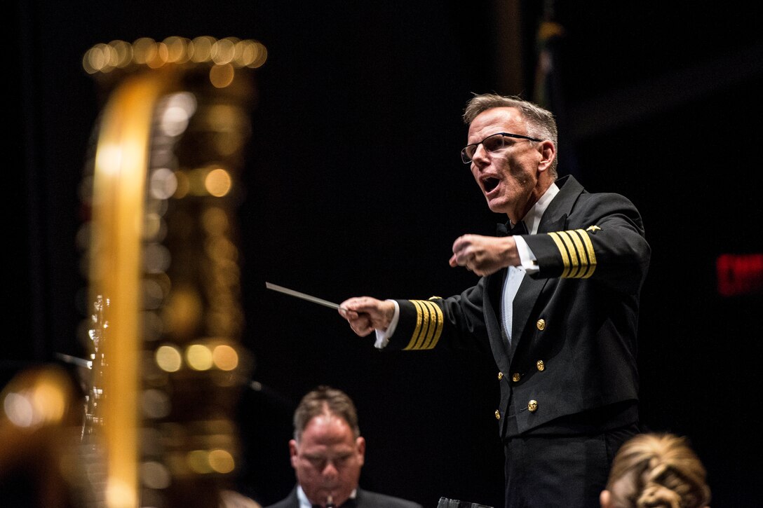 A conductor stands and gestures with his baton as musicians play instruments.