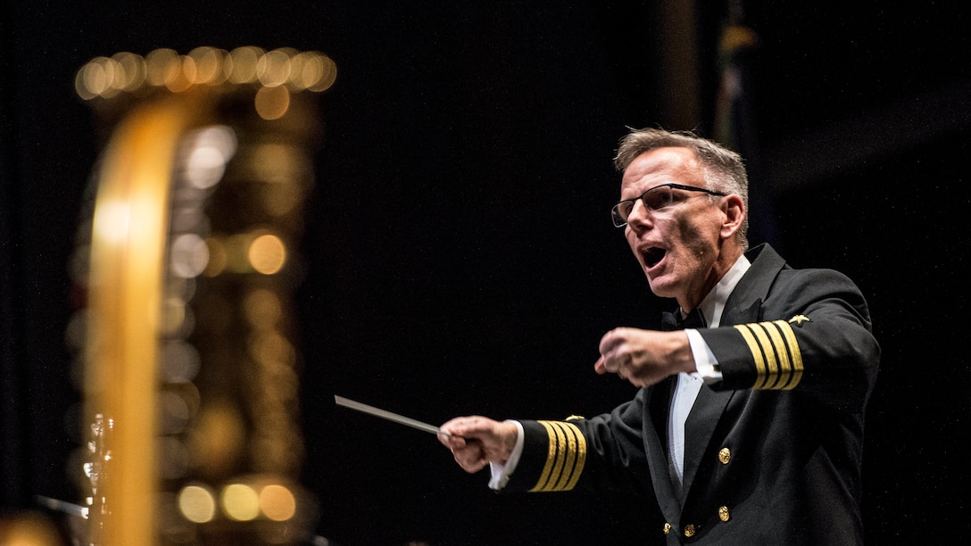 A conductor stands and gestures with his baton as musicians play instruments.