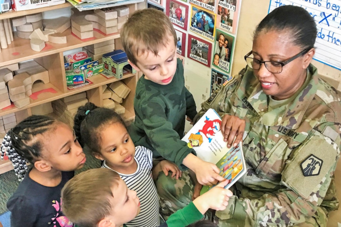 Children gather around a soldier reading to them, and study and touch the book she's holding.