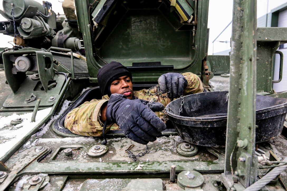 A soldier works on parts on top of a vehicle's exterior while perched partway outside the top opening of the vehicle.