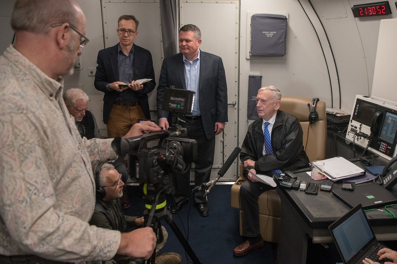 Defense secretary speaks with reporters in aircraft cabin.