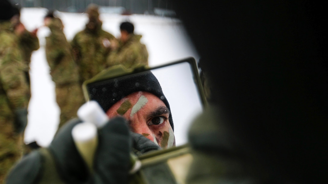 A soldier, whose face is visible in a hand-held mirror, applies camouflage paint in a snowy field with soldiers gathered nearby.