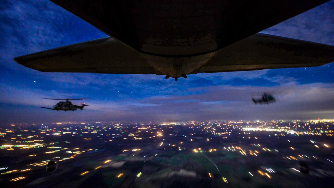 Two helicopters trail an airplane's tail in a sapphire-blue sky over city lights.