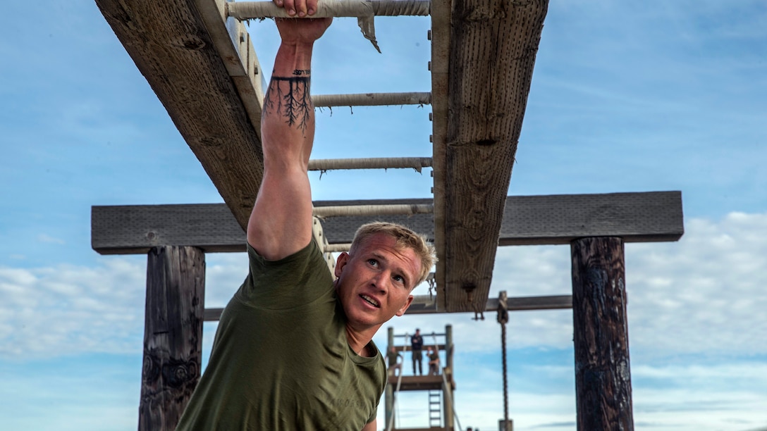 A sailor hangs from one arm on a bar obstacle.