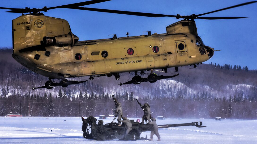 Two soldiers standing on a howitzer reach up to connect slings to a hovering helicopter as snow gusts around them.