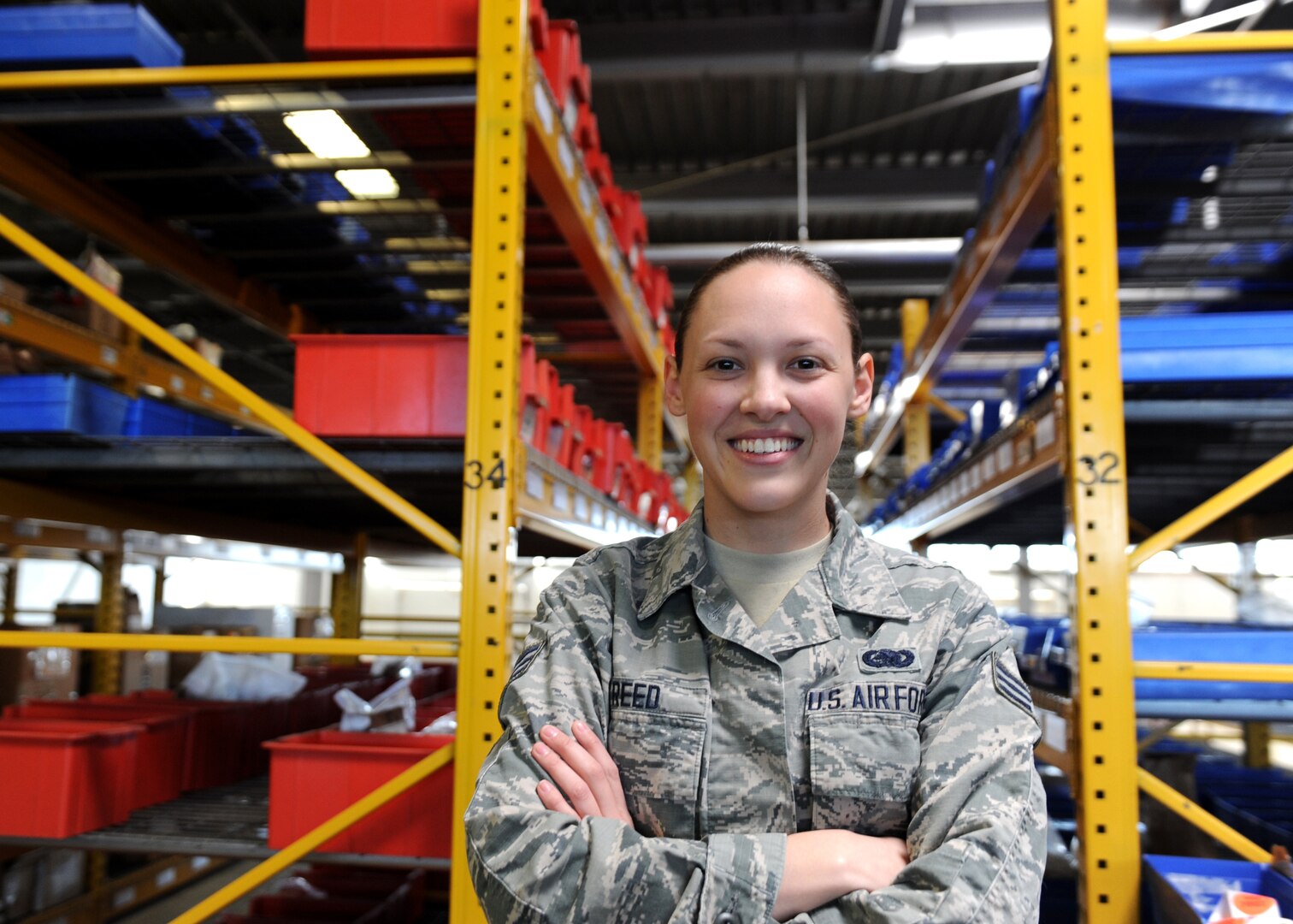 A smiling female stands in front of yellow shelves housing aircraft parts in a large warehouse.