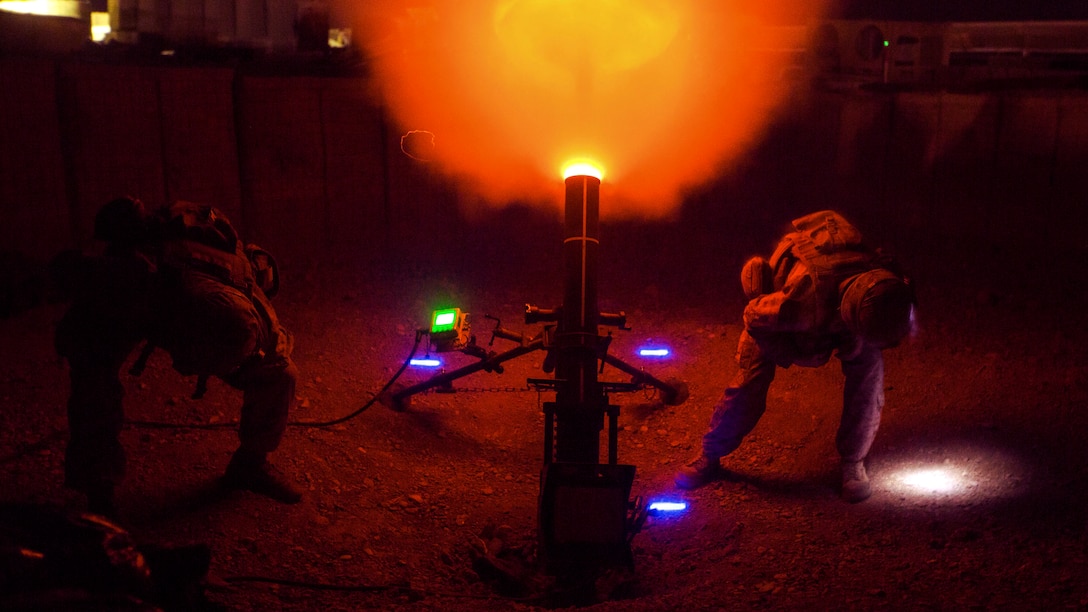 Orange flames burst from a howitzer, illuminating a night sky, as Marines crouch on either side of the weapon.