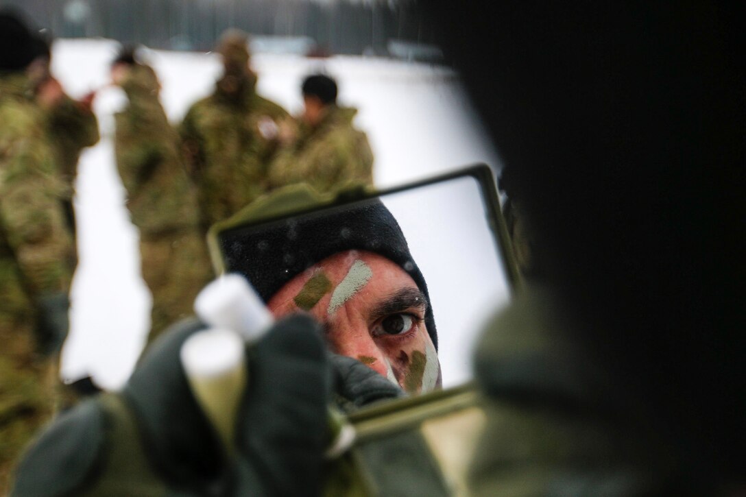 A soldier, whose face is visible in a hand-held mirror, applies camouflage paint in a snowy field with soldiers gathered nearby.