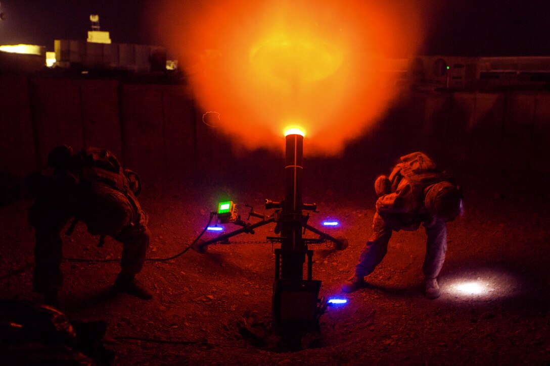 Orange flames burst from a howitzer, illuminating a night sky, as Marines crouch on either side of the weapon.