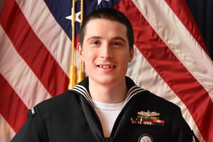 A sailor head shot in front of a flag.