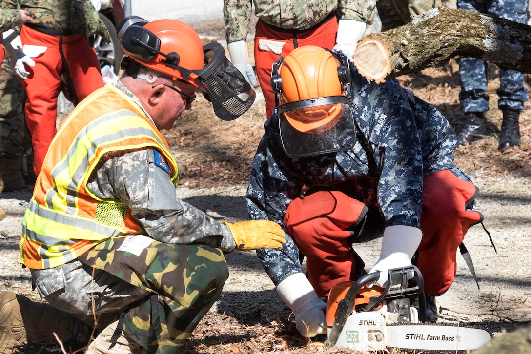A soldier instructs a sailor on the safety and chainsaw operation.