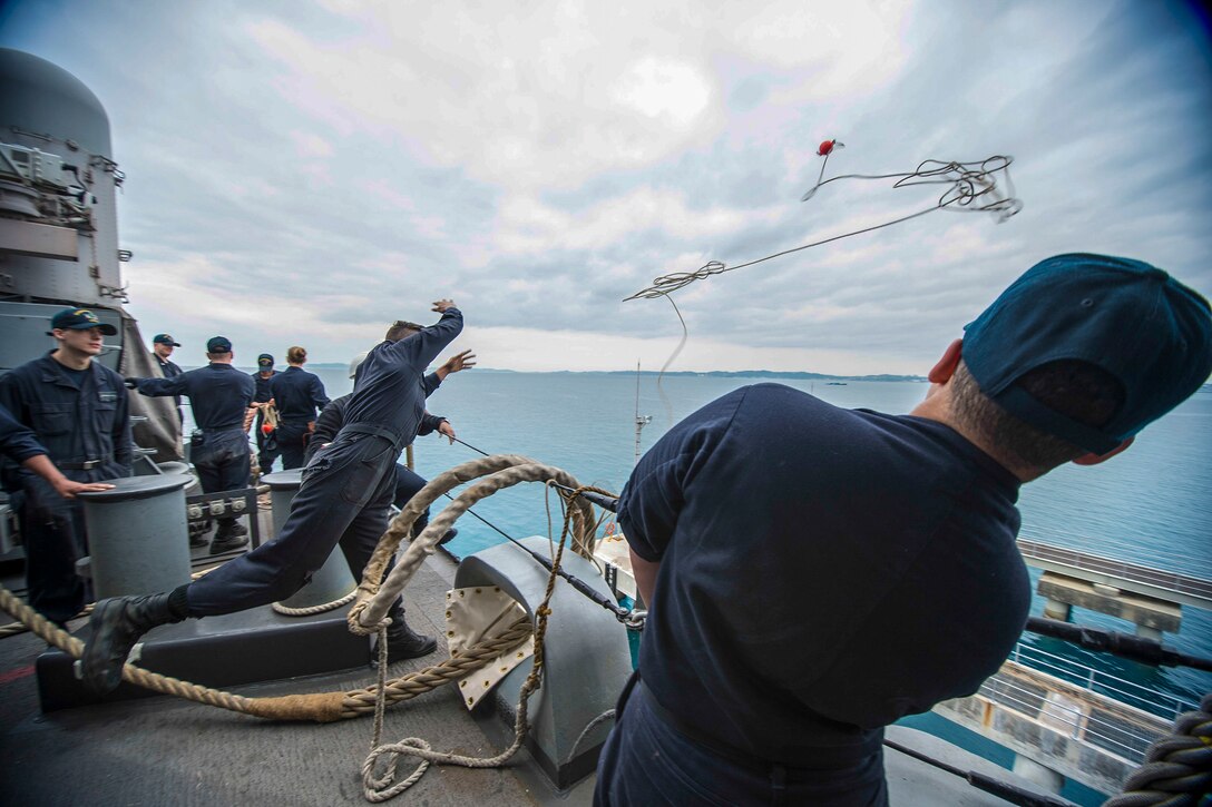 A sailor throws a messenger line from a ship to sailors on a pier.