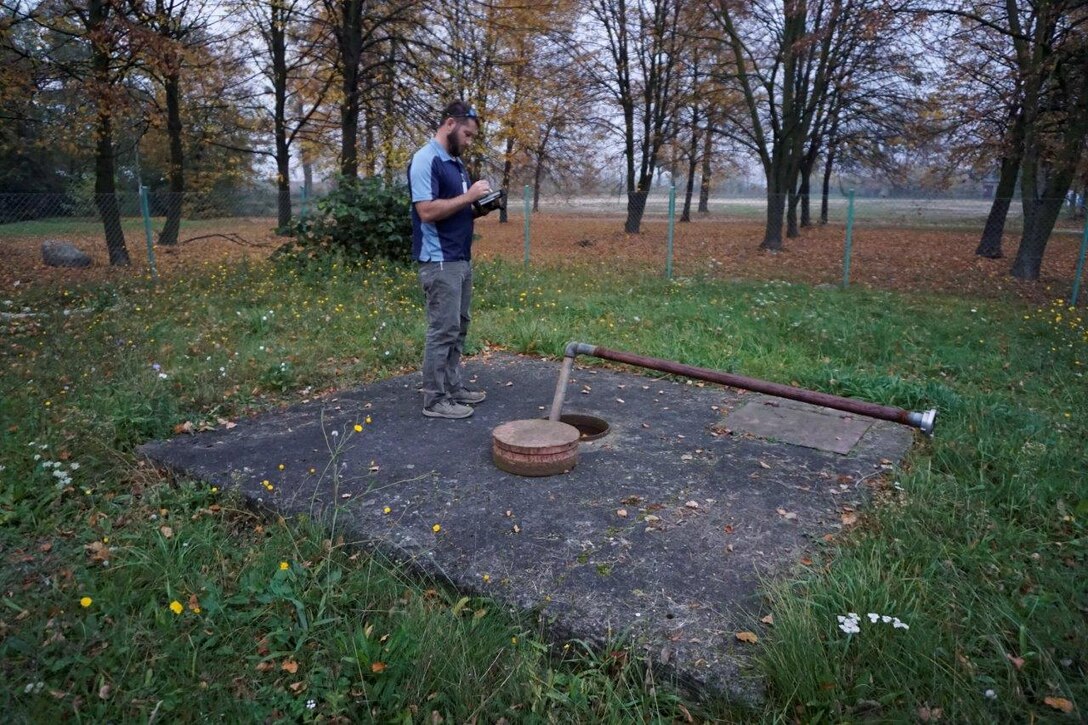 A man stands on a concrete block, looking into a manhole.
