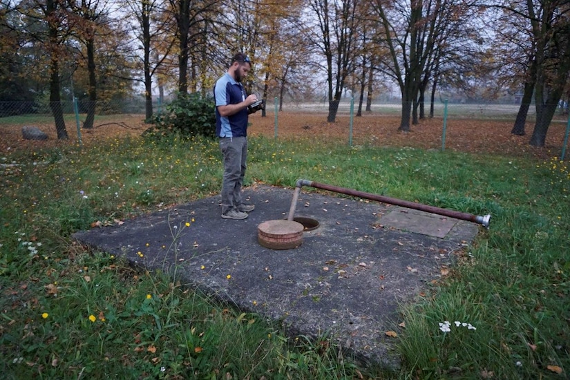 A man stands on a concrete block, looking into a manhole.