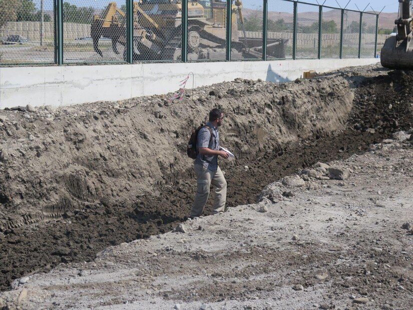 A man stands in a trench, with a large bulldozer in the background.