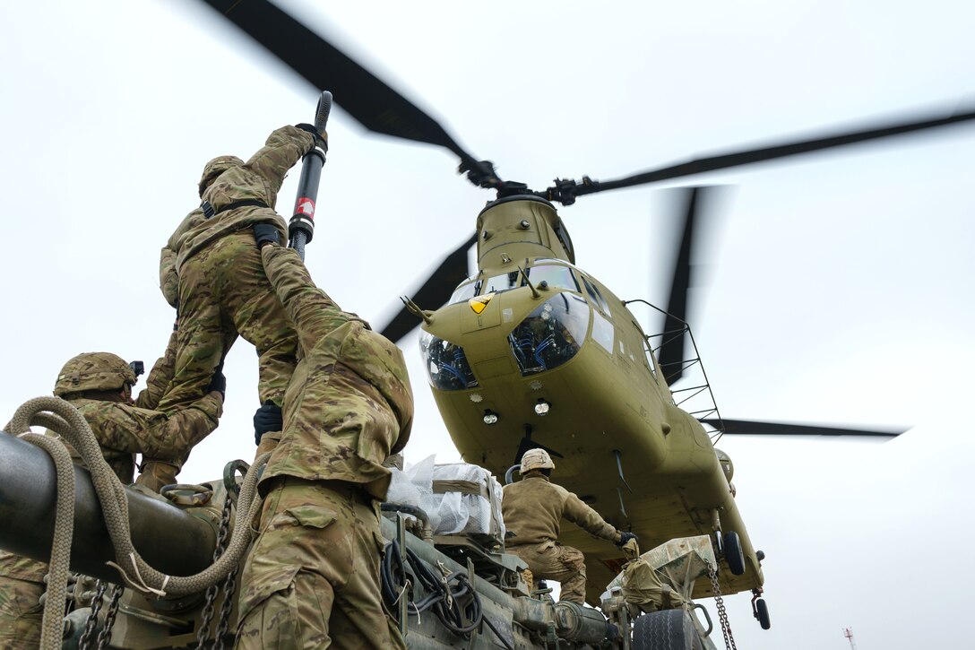 Soldiers reach up to hook a M777 howitzer to a CH-47 Chinook helicopter