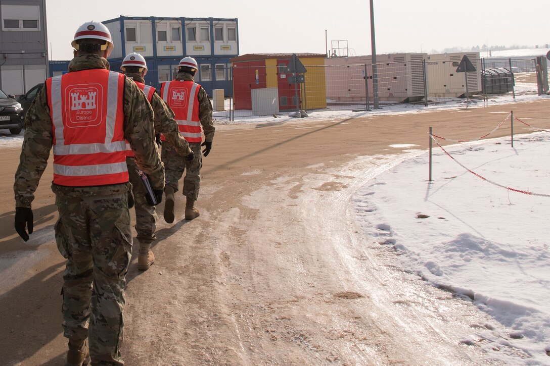 Three men in military uniforms walk along a dirt path on a construction site.