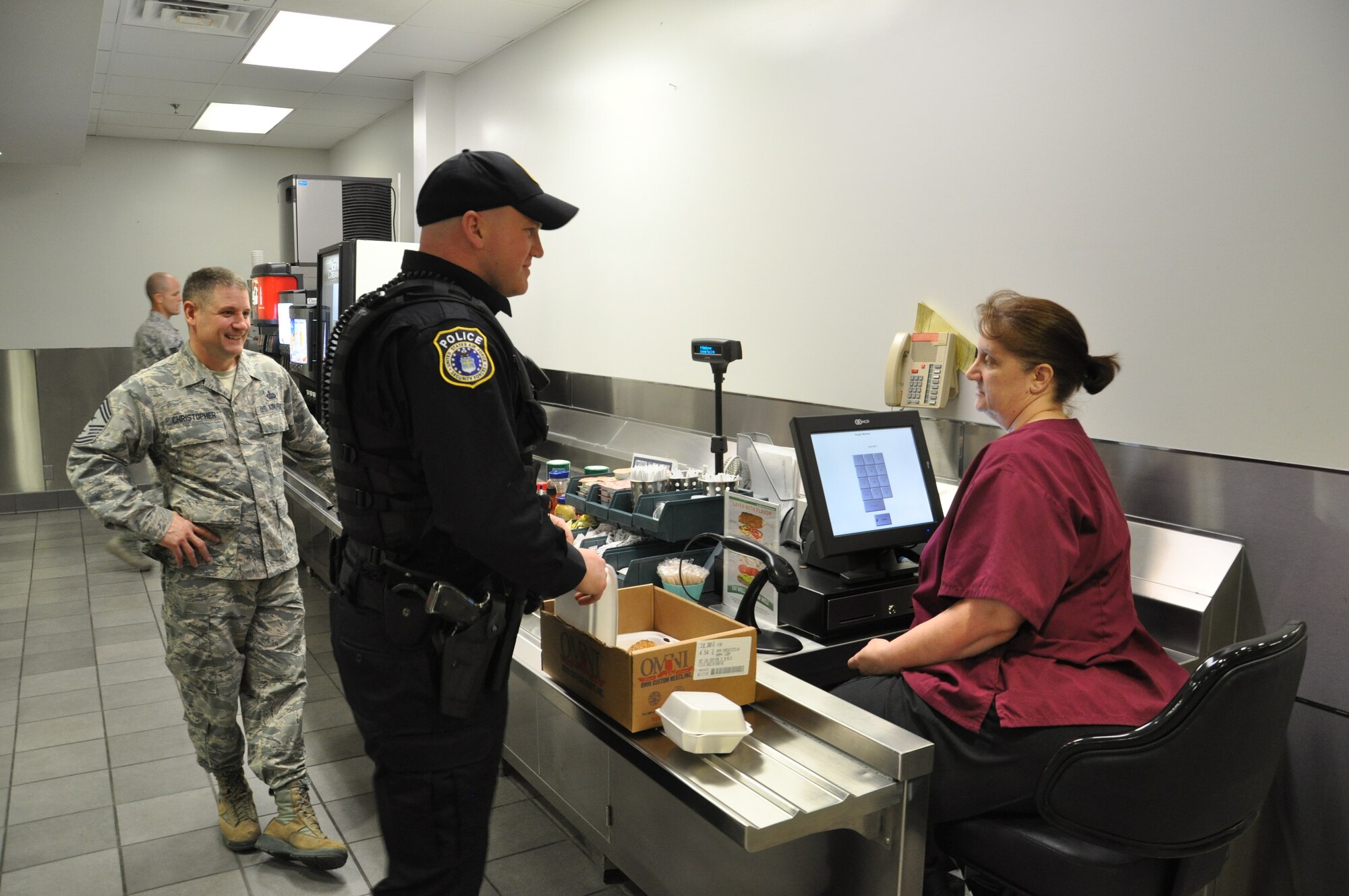 Nancy Williams, Pitsenbarger Dining Facility cashier, rings up Officer Ryan Gray’s main entrée “to go” while Chief Master Sgt. Thomas Christopher looks on at the Pitsenbarger Dining Facility located in Building 1214, Area A, Kittyhawk. (U.S. Air Force photo/W. Eugene Barnett Jr.)