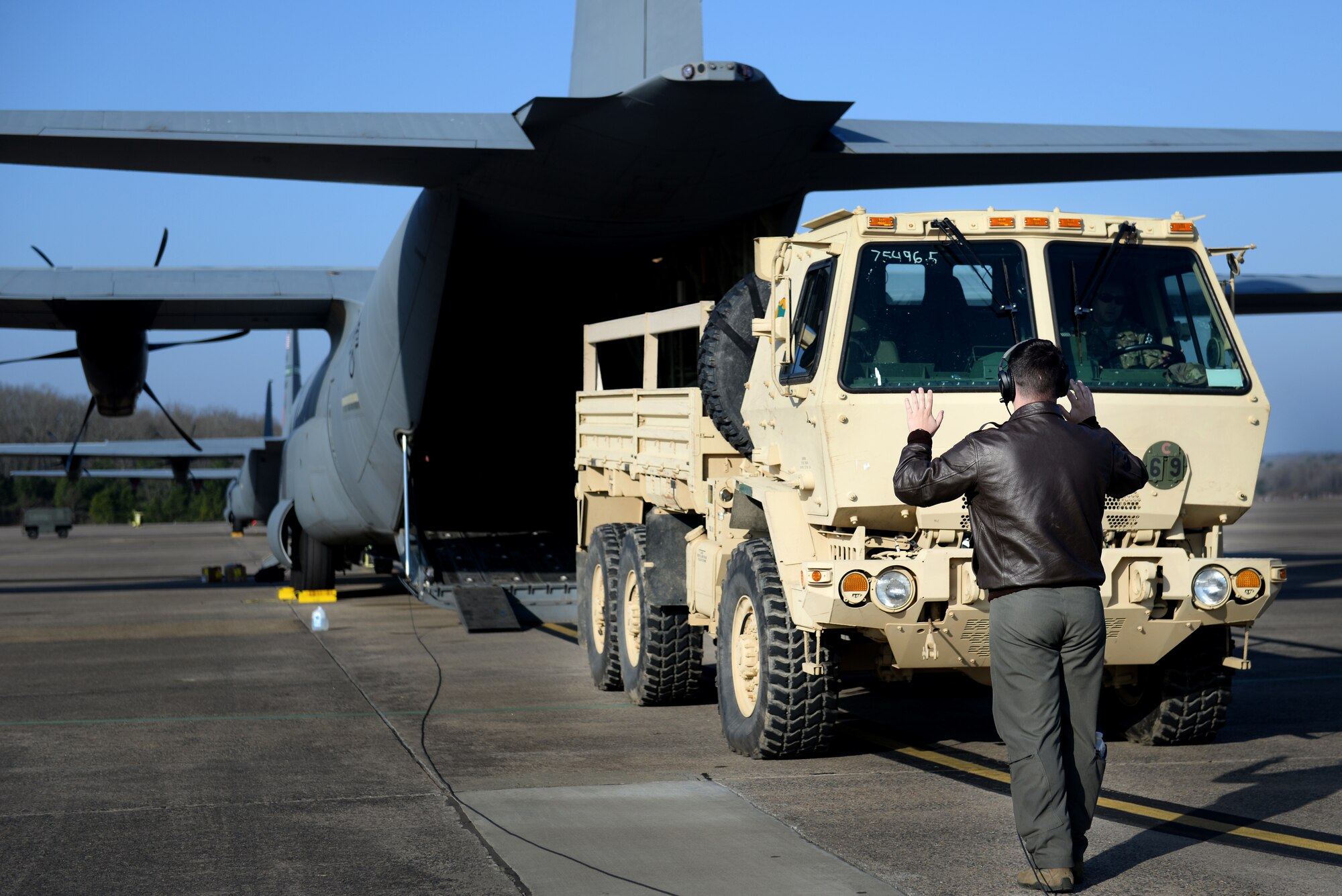 An Airman wearing a flight suit and leather jacket directs a medium tactical vehicle driver into a C-130 J.