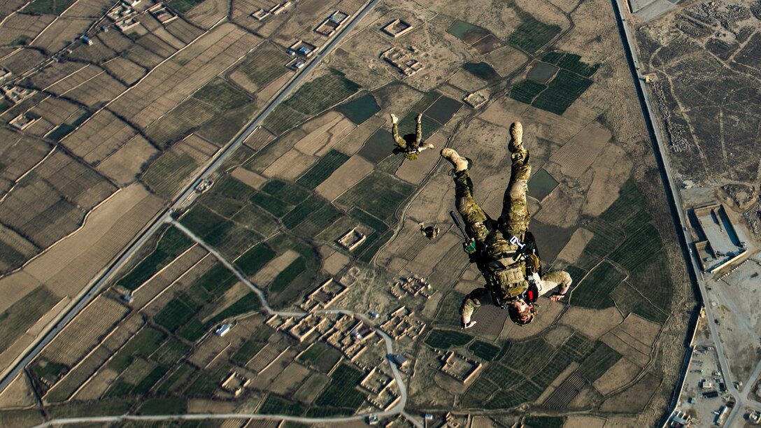 Three airmen free-fall with their limbs splayed before their parachutes have opened over brown and green terrain.