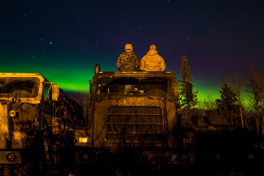 Two Marines, shown from behind, sit atop a truck and look at the northern lights.