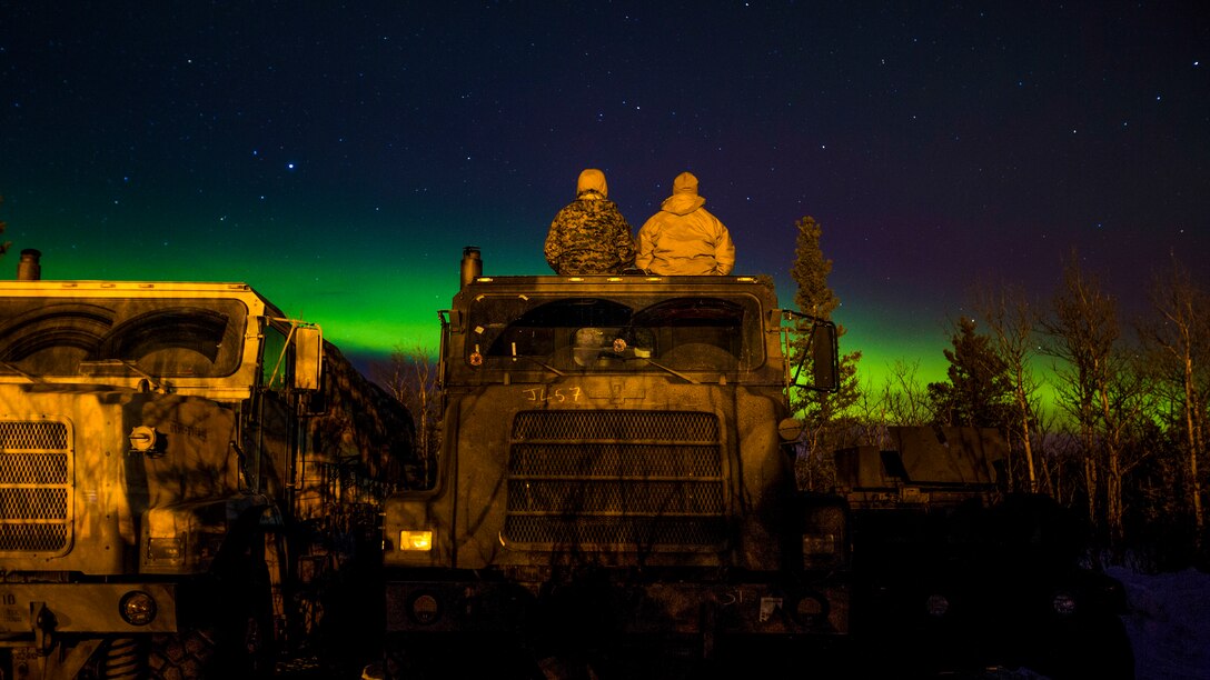 Two Marines, shown from behind, sit atop a truck and look at the northern lights.